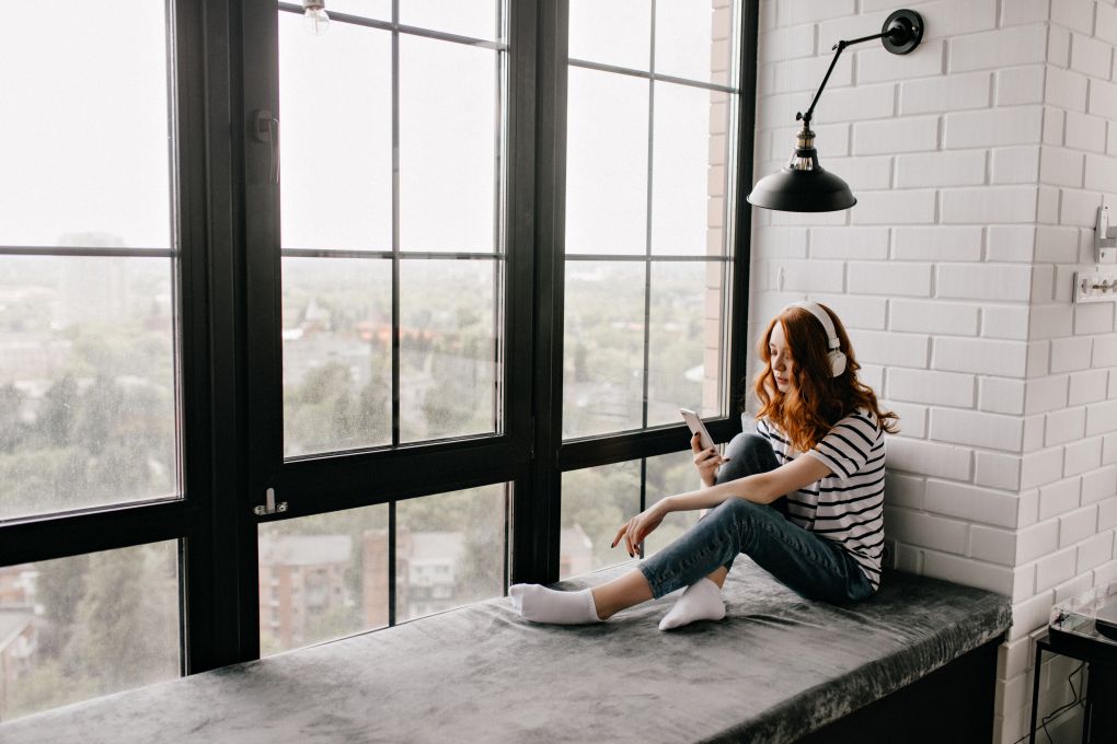 Red-haired girl sitting by a sunny window with headphones, enjoying UV-protected indoor comfort